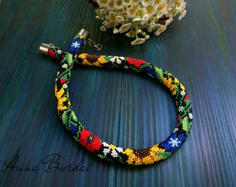 May include: A colorful beaded necklace with a floral pattern. The necklace is made of black, red, yellow, green, and blue beads. The necklace is on a blue wooden surface.