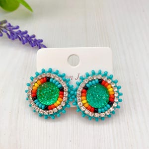 Könnte beinhalten: Ein Paar türkisfarbene Perlenohrringe. Jeder Ohrring hat einen zentralen grünen Glitzerkreis, umgeben von einem Ring aus farbigen Perlen in Rot, Orange, Gelb und Schwarz. Der äußere Rand ist mit klaren Strasssteinen verziert.