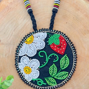 May include: A beaded necklace with a circular pendant displaying a floral and fruit design. The pendant features white daisies with yellow centres and a red strawberry with green leaves, set against a black backdrop. The necklace has a black braided cord.
