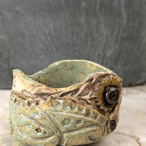 May include: A small, hand-built ceramic bowl with a light green glaze and dark brown speckles. The exterior features intricate, raised relief patterns and two dark, circular bead-like accents. The bowl rests on three small, rounded feet.