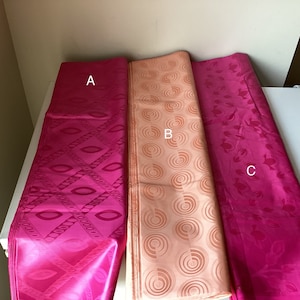 May include: Three rolls of patterned fabric in shades of pink and peach. The fabric on the left has a diamond and oval design, the middle fabric has a circular pattern, and the fabric on the right has a floral design.