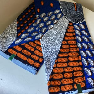 May include: Folded fabric with a vibrant patchwork design. The fabric features a mix of blue, orange, white, and black patterns, including oval shapes, brick-like designs, and swirling motifs. The fabric is labeled "Veritable Wax".