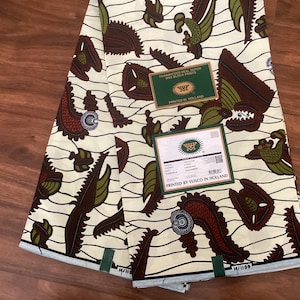 May include: Cream-colored fabric with a repeating pattern of brown and green stylized shapes. The fabric has a wavy design and includes two green labels with text that reads "GUARANTEED REAL DUTCH WAX BLOCK PRINTS PRINTED IN HOLLAND".