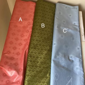 May include: Three rolls of fabric in shades of coral, olive green, and light blue. The coral fabric has a diamond pattern, the green has a square pattern, and the blue has a floral design. The fabrics are likely for sewing or crafting.