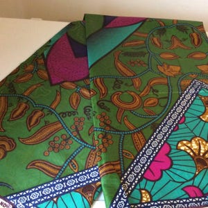 May include: Folded green fabric with a vibrant floral and leaf pattern in shades of brown, teal, and pink. The fabric has a detailed border with geometric designs and a teal and white trim.
