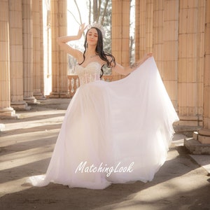 May include: A woman wearing a white wedding dress with a lace bodice and a flowing skirt. She is standing in a colonnade with sunlight streaming in.