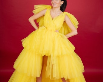 May include: A woman wearing a bright yellow tulle dress with a high-low hemline. The dress has a V-neckline and ruffled sleeves. She is standing on a red background.