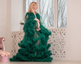May include: A pregnant person wearing a long, green, tulle gown with ruffles. The gown is sheer and has a flowing silhouette. The person is standing in front of a window with a white, decorative lattice.