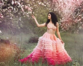 May include: A woman in a pink and white ombre tulle dress stands in a field of blooming trees. The trees are in full bloom with white flowers and the woman is reaching out to touch a branch.