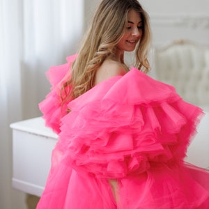 May include: A woman wearing a bright pink tulle dress with multiple layers of ruffles. The dress is short and has a loose, flowing fit. The woman is standing in front of a white piano.