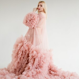 Pu&ograve; includere: Una donna indossa un abito di tulle rosa con un ampio bordo di volant. L'abito &egrave; trasparente e ha una silhouette fluida.