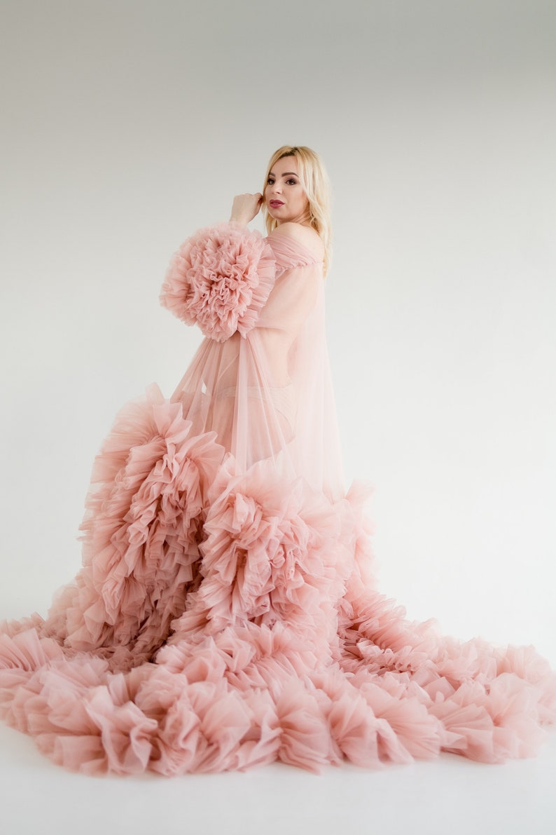Pu&ograve; includere: Una donna indossa un abito di tulle rosa con un ampio bordo di volant. L'abito &egrave; trasparente e ha una silhouette fluida.