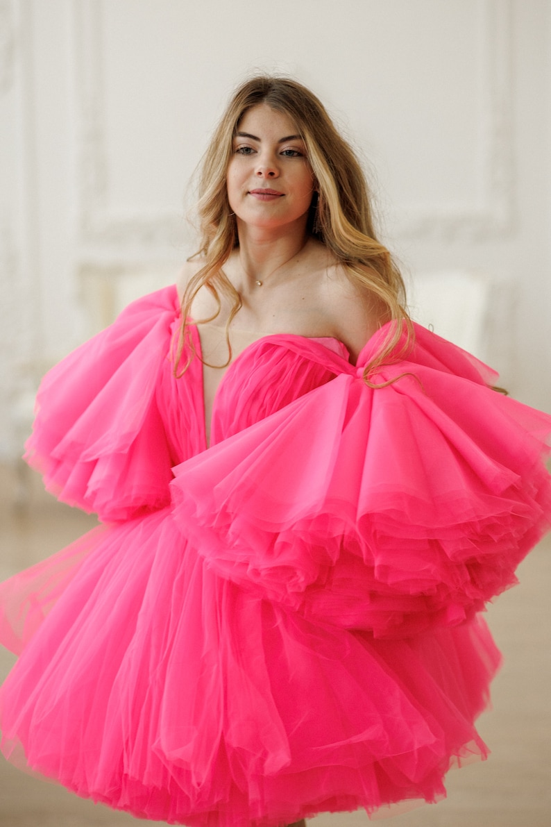 May include: A woman wearing a bright pink tulle dress with large, ruffled sleeves. The dress is strapless and has a sweetheart neckline.