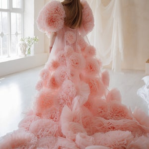 May include: A light pink gown with large, ruffled floral accents. The dress features voluminous, flower-like sleeves and a long, flowing train. The gown is made of a soft, sheer fabric, and is suitable for special occasions.