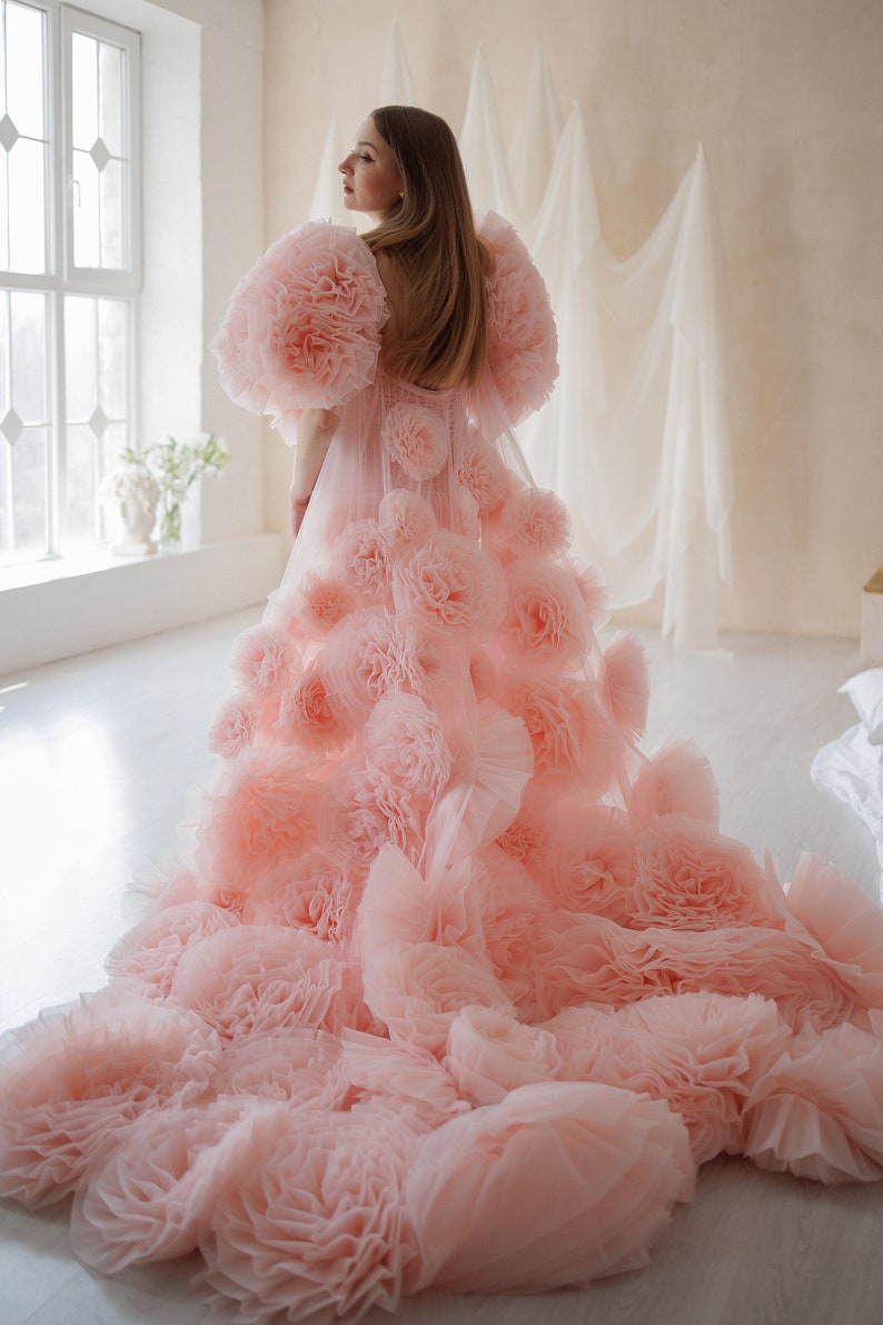 May include: A light pink gown with large, ruffled floral accents. The dress features voluminous, flower-like sleeves and a long, flowing train. The gown is made of a soft, sheer fabric, and is suitable for special occasions.