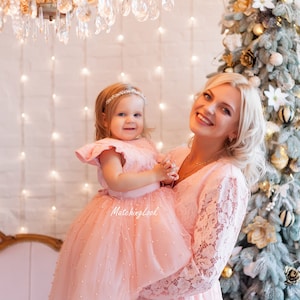 Puede incluir: Una madre y su hija visten vestidos de encaje rosa a juego con adornos de perlas. La ni&ntilde;a lleva un peque&ntilde;o ramo de flores. Est&aacute;n de pie frente a un &aacute;rbol de Navidad decorado con adornos dorados y blancos.