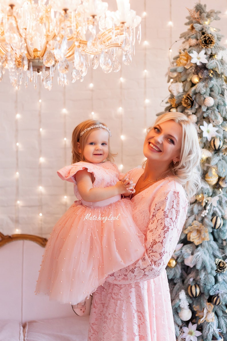 Puede incluir: Una madre y su hija visten vestidos de encaje rosa a juego con adornos de perlas. La ni&ntilde;a lleva un peque&ntilde;o ramo de flores. Est&aacute;n de pie frente a un &aacute;rbol de Navidad decorado con adornos dorados y blancos.