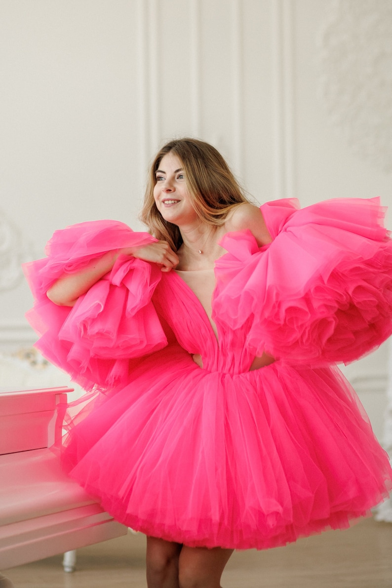 May include: A woman wearing a bright pink tulle dress with large, ruffled sleeves. The dress is short and has a sweetheart neckline. The woman is standing in front of a white piano.