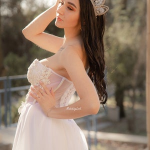 May include: A woman wearing a white strapless gown with a tulle skirt and a silver crown with rhinestones. The gown has lace detailing and the woman is holding her hand to her head.