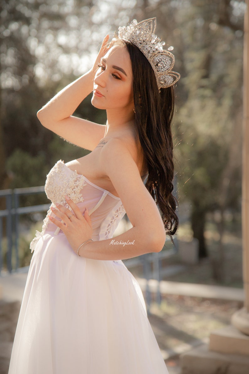 May include: A woman wearing a white strapless gown with a tulle skirt and a silver crown with rhinestones. The gown has lace detailing and the woman is holding her hand to her head.