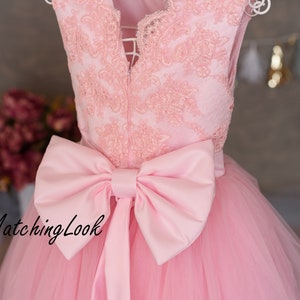 Puede incluir: Un vestido de tul rosa con un corpi&ntilde;o de encaje y un gran lazo de sat&eacute;n rosa en la cintura. El vestido tiene un escote en V y es sin mangas. El vestido est&aacute; en una percha y el fondo es una pared gris claro.