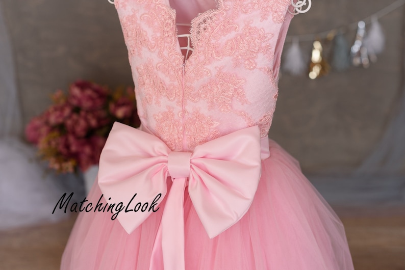 Puede incluir: Un vestido de tul rosa con un corpi&ntilde;o de encaje y un gran lazo de sat&eacute;n rosa en la cintura. El vestido tiene un escote en V y es sin mangas. El vestido est&aacute; en una percha y el fondo es una pared gris claro.