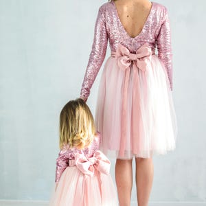 May include: A mother and daughter wearing matching pink sequin and tulle dresses with large pink bows. The dresses have long sleeves and a V-shaped back.