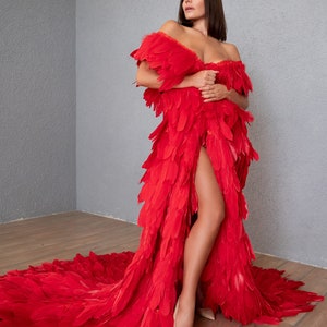May include: A woman wearing a long, red feather dress with a thigh-high slit. The dress has a flowing train and is made of many layers of feathers.