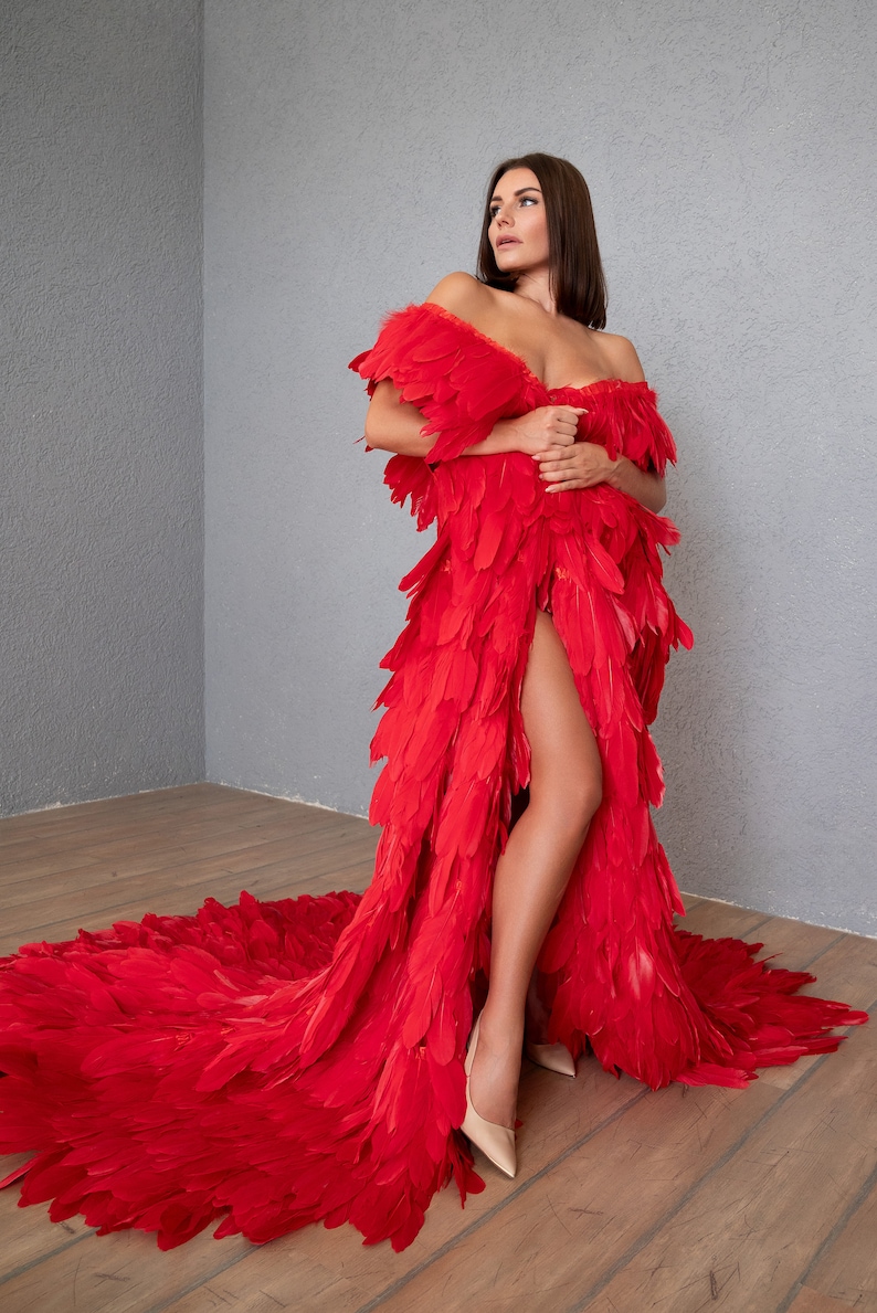 May include: A woman wearing a long, red feather dress with a thigh-high slit. The dress has a flowing train and is made of many layers of feathers.