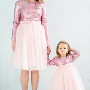 May include: A mother and daughter wearing matching pink sequin tops and tulle skirts. The tops have long sleeves and the skirts are knee-length.