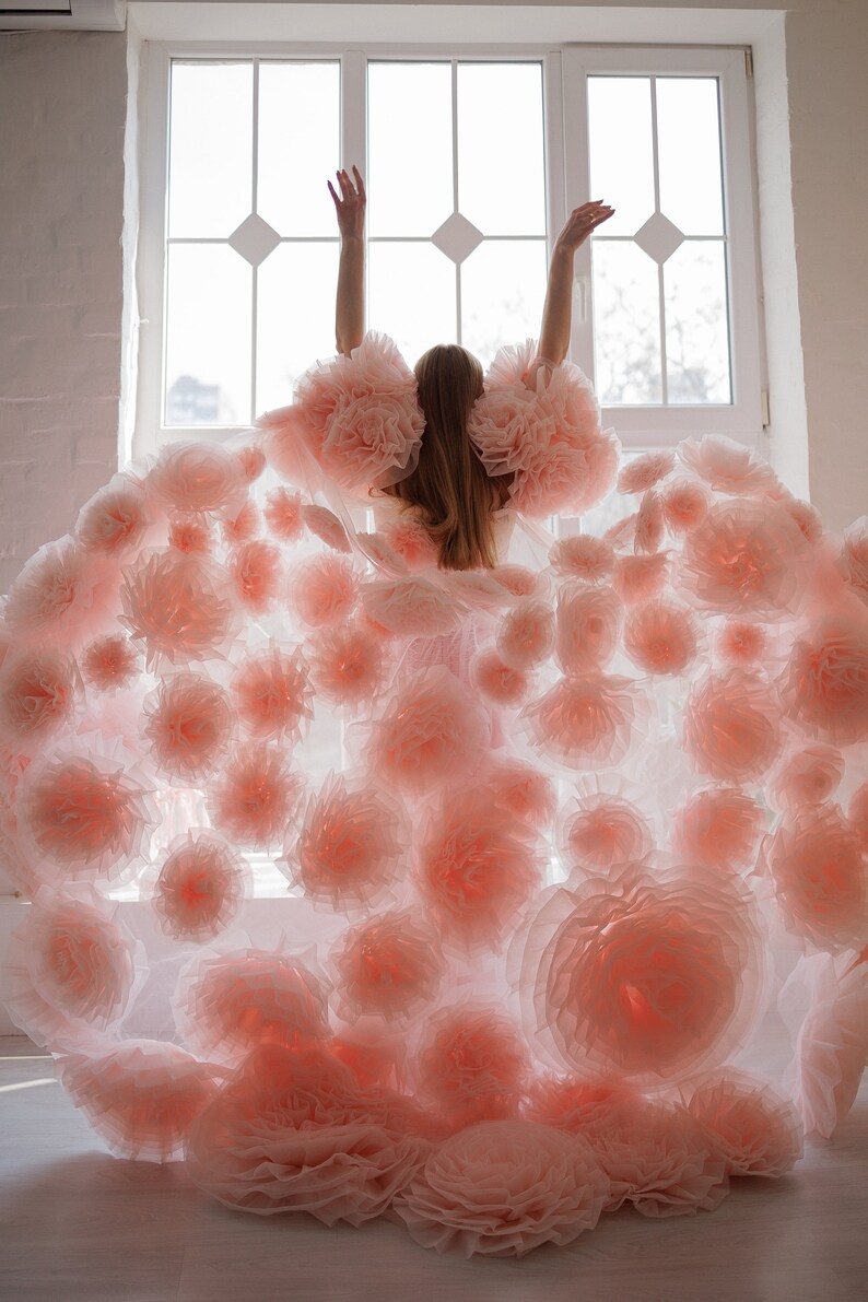 May include: A person seen from the back, wearing a light pink dress adorned with numerous large, soft, flower-like embellishments. The dress has voluminous sleeves and a flowing skirt, creating a dramatic silhouette. The person's arms are raised.