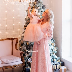 Puede incluir: Una madre y su hija llevan vestidos de encaje rosa a juego y est&aacute;n de pie frente a un &aacute;rbol de Navidad decorado. La hija est&aacute; siendo sostenida por su madre y est&aacute; alcanzando un candelabro.