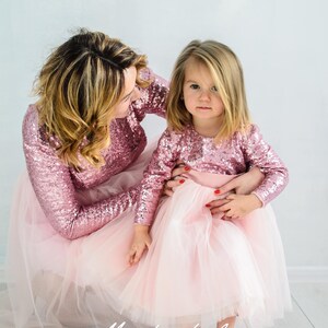 May include: A mother and daughter wearing matching pink sequin long-sleeved tops and pink tulle skirts. The girl is sitting on the floor with her mother's arm around her. The text "Matching Look" is visible in the bottom center of the image.