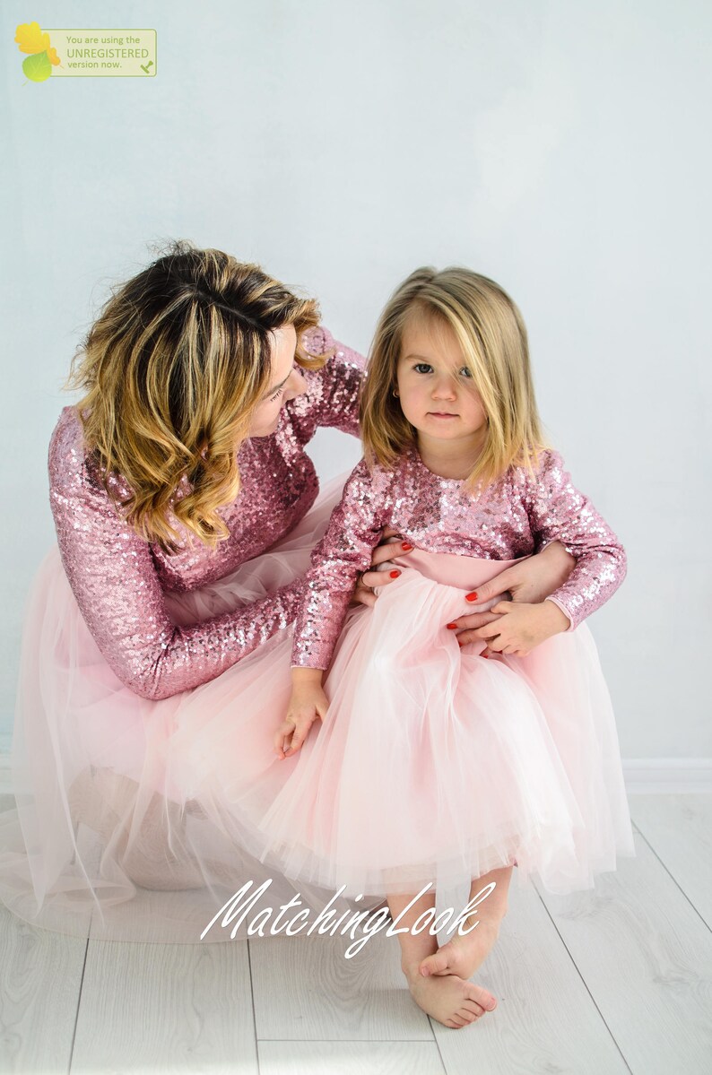 May include: A mother and daughter wearing matching pink sequin long-sleeved tops and pink tulle skirts. The girl is sitting on the floor with her mother's arm around her. The text "Matching Look" is visible in the bottom center of the image.