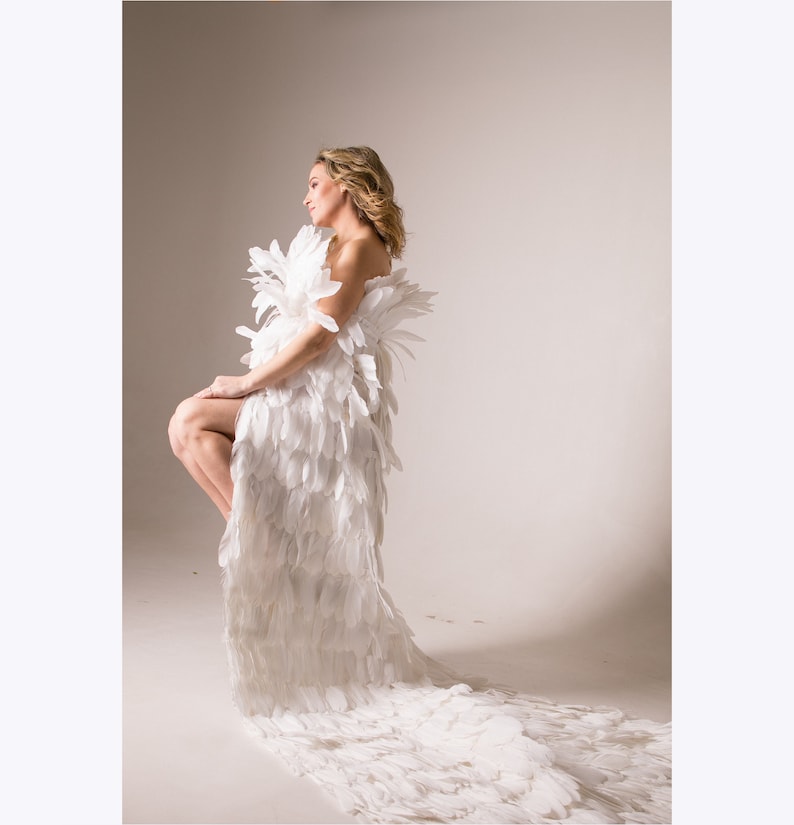 May include: A woman wearing a white feather dress with a long train. The dress is made of many individual feathers that are arranged in a cascading pattern. The woman is standing in a studio setting with a white background.