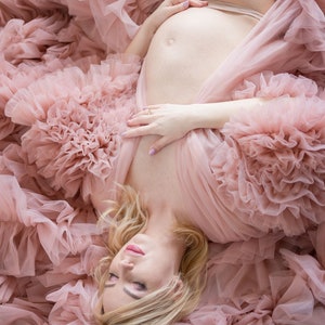 Pu&ograve; includere: Una donna incinta che indossa un abito di tulle rosa con maniche a sbuffo. &Egrave; sdraiata sulla schiena con le mani appoggiate sulla pancia.