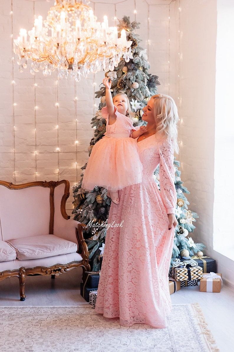 Puede incluir: Una madre y su hija llevan vestidos de encaje rosa a juego y est&aacute;n de pie frente a un &aacute;rbol de Navidad. La hija est&aacute; siendo sostenida por su madre y est&aacute; alcanzando un candelabro. El texto "Matching Look" es visible en la imagen.