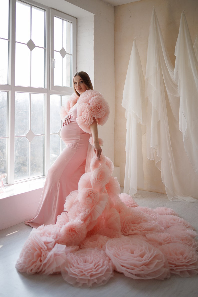 May include: A light pink maternity gown with a long train of large, ruffled fabric flowers. The gown features a fitted silhouette and a dramatic, floral-adorned shoulder detail. The model is standing near a window, with sheer white curtains in the background.