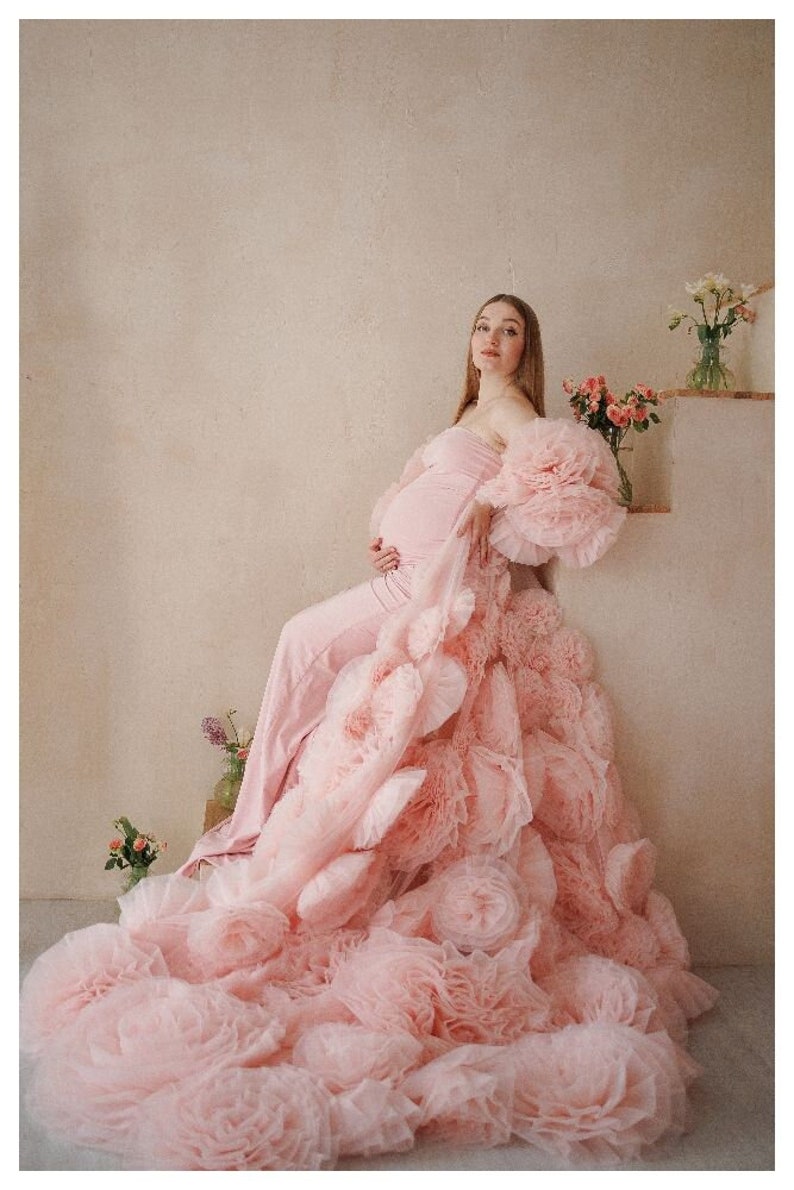 May include: A light pink maternity gown with a strapless bodice and a flowing skirt. The gown features large, ruffled sleeves and a cascade of fabric that creates a voluminous effect. The model is seated, with flowers in the background.