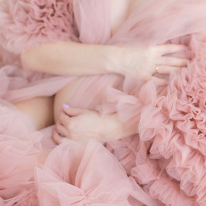 Pu&ograve; includere: Una donna indossa un abito di tulle rosa con volant. L'abito &egrave; molto fluido e ha molti strati. La donna tiene le mani sulla pancia.