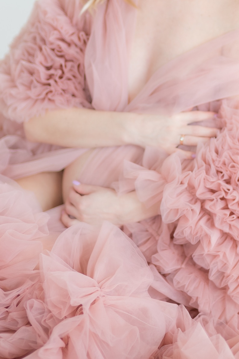Pu&ograve; includere: Una donna indossa un abito di tulle rosa con volant. L'abito &egrave; molto fluido e ha molti strati. La donna tiene le mani sulla pancia.