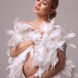 May include: A pregnant woman wearing a white feather boa. The boa is draped over her shoulders and covers her chest and stomach. She is wearing a silver earring.