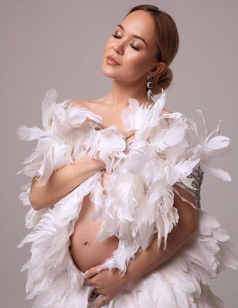 May include: A pregnant woman wearing a white feather boa. The boa is draped over her shoulders and covers her chest and stomach. She is wearing a silver earring.