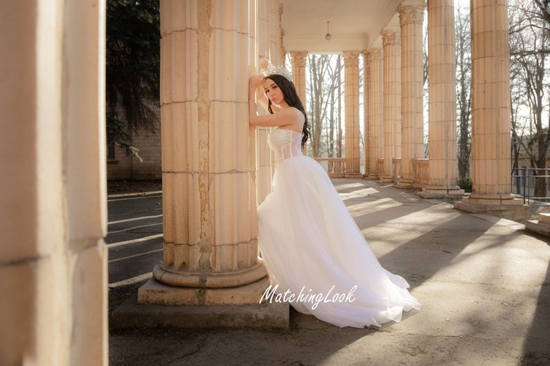 May include: A woman in a white wedding dress with a tulle skirt stands in front of a column. She is wearing a tiara and has her hair pulled back. The background is a colonnade with trees visible in the distance.