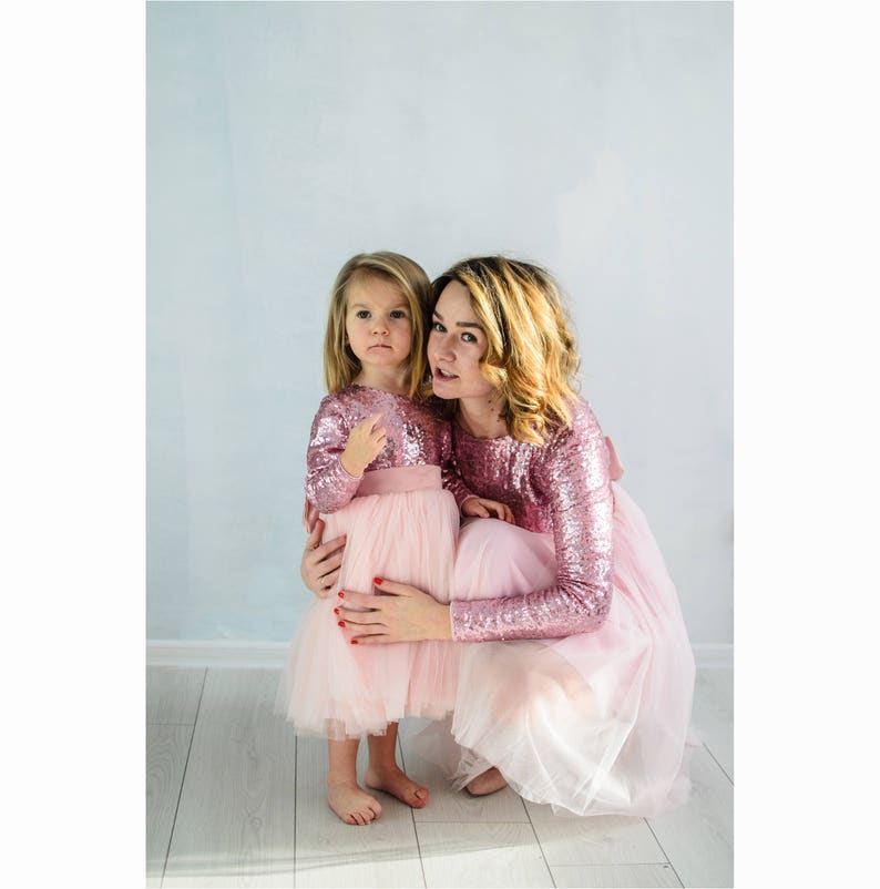 May include: A mother and daughter wearing matching pink sequin dresses with tulle skirts. The dresses have long sleeves and a pink ribbon at the waist.