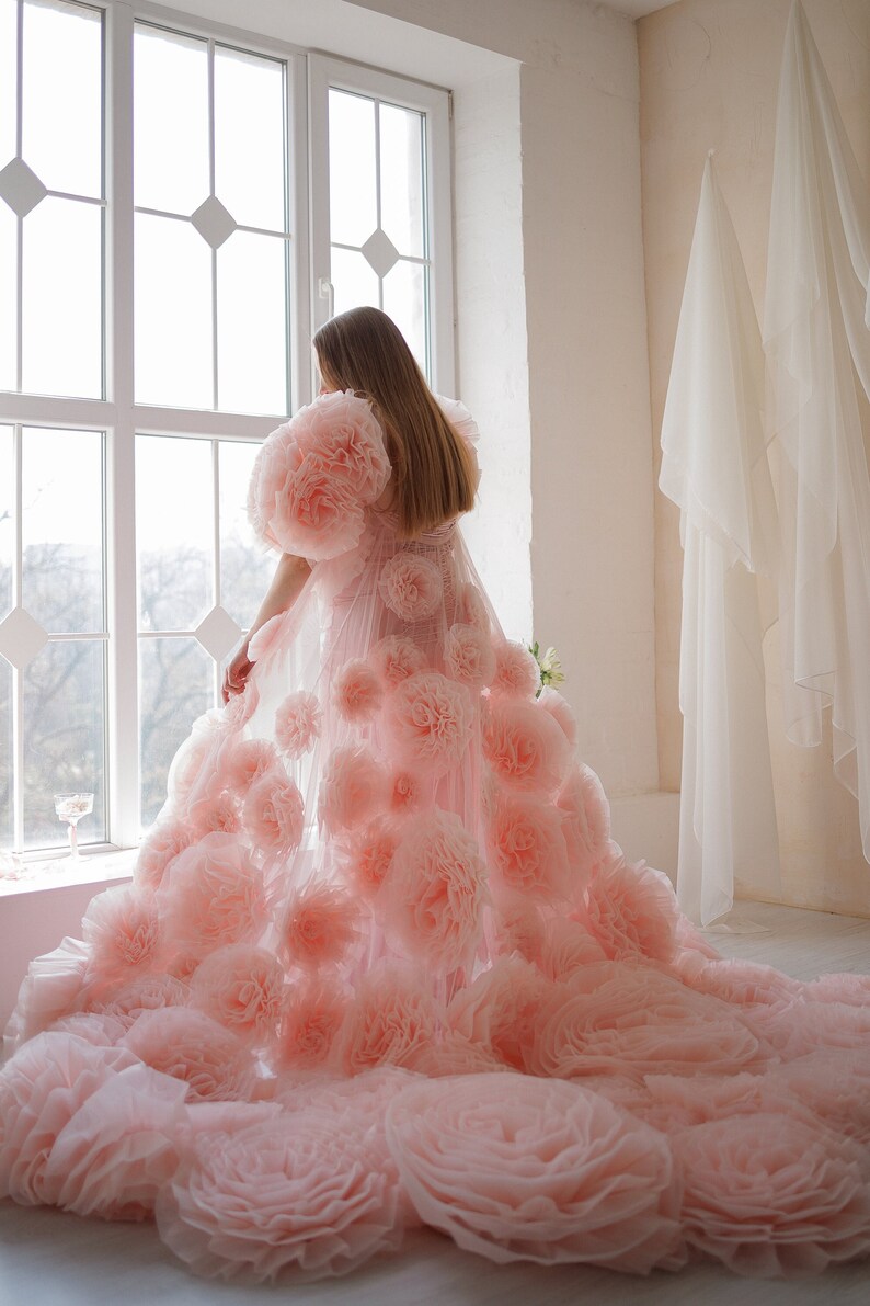 May include: A light pink gown with large, fluffy floral embellishments. The dress features voluminous, rose-like details cascading down the skirt and on the shoulders. The gown is made of a sheer fabric, creating a dreamy, ethereal look.