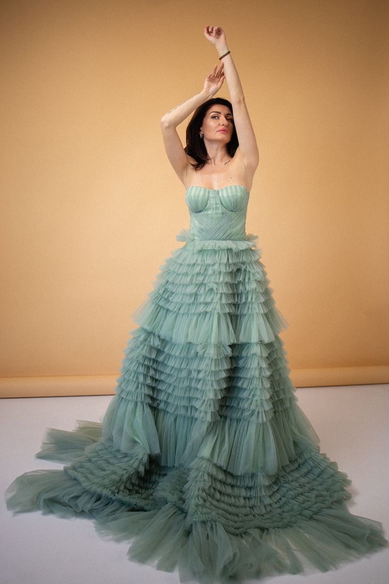Pu&ograve; includere: Una donna indossa un abito di tulle verde chiaro con pi&ugrave; strati di balze. L'abito ha un corpetto senza spalline con un design aderente. La donna &egrave; in piedi con il braccio alzato sopra la testa.