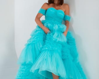May include: A turquoise off-the-shoulder gown with a layered tulle skirt. The dress features a sweetheart neckline and short sleeves. The model is wearing nude-colored heels.