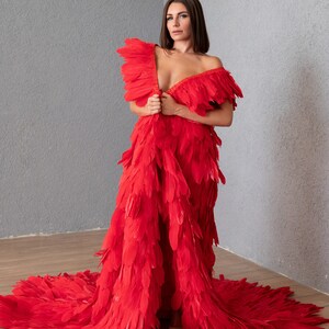 May include: A woman wearing a red feather dress with a long train. The dress is made of many layers of feathers and has a deep V-neckline.