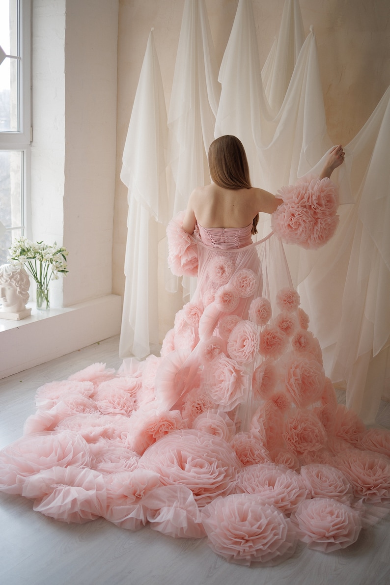 May include: A strapless pink gown with a corset-style bodice and a flowing skirt adorned with large, ruffled fabric flowers. The sleeves are also embellished with the same floral detail. The gown is made of a soft, sheer material.